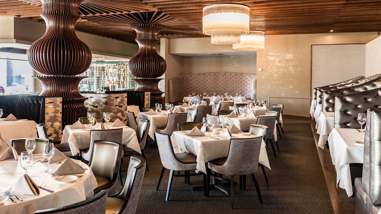 The dining room at Dee Lincoln Prime, with bronze-colored, rounded columns that blend into a ceiling of narrow brown slats; gray cloth and leather chairs sit at tables with wineglasses, and a wall of wine bottles can be seen in the background; contemporary-design chandeliers hang above